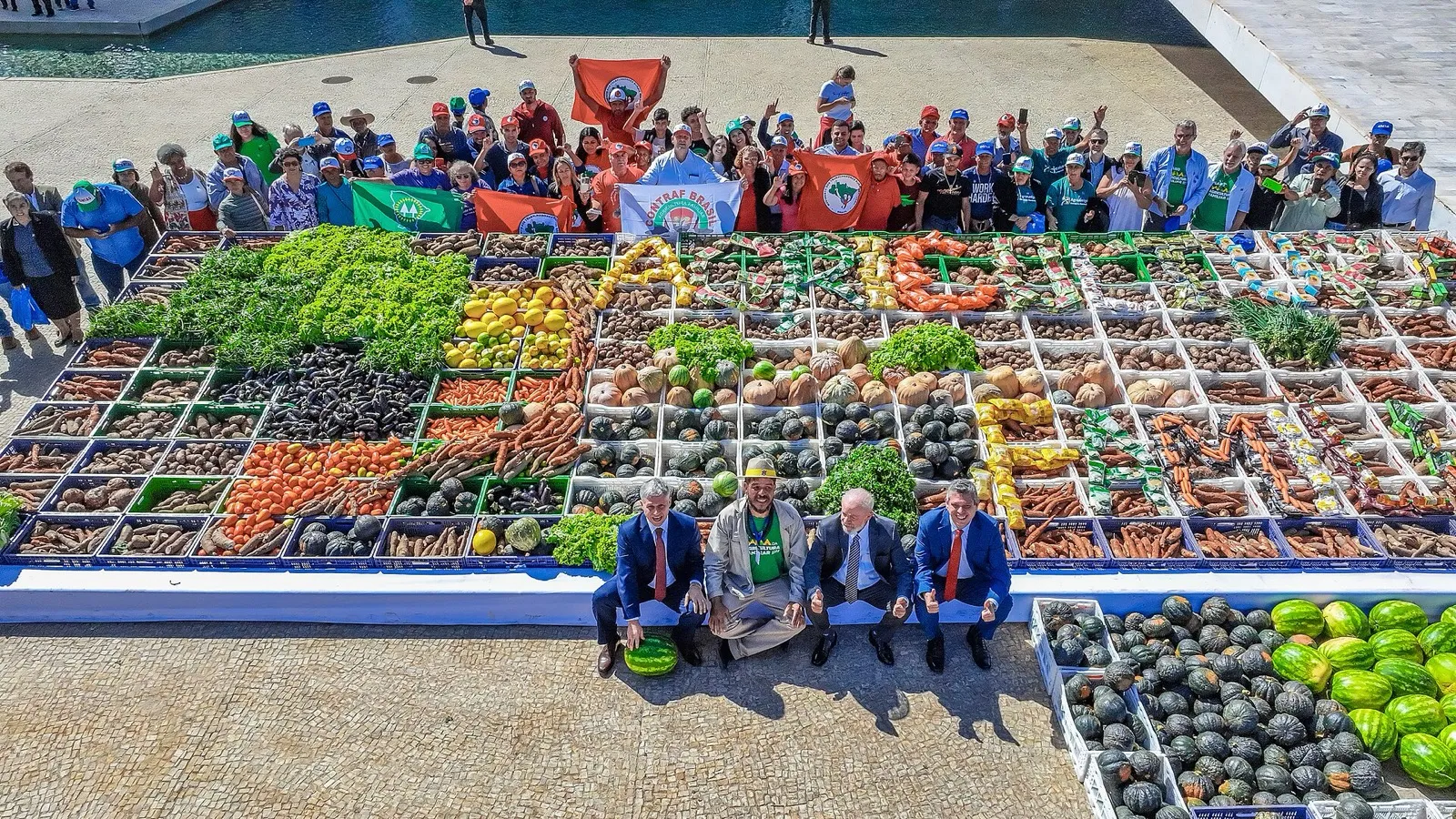 Quatro homens sentados diante de um grande mosaico de legumes e verduras, com agricultores segurando bandeiras de movimentos rurais ao fundo