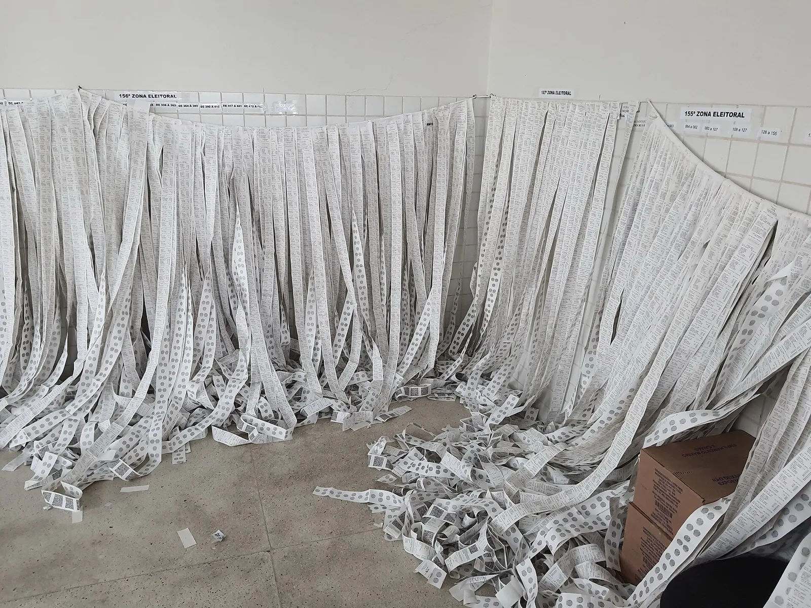 Tiras de boletins de urna penduradas em uma parede, com placas da 155ª e da 156ª zonas eleitorais