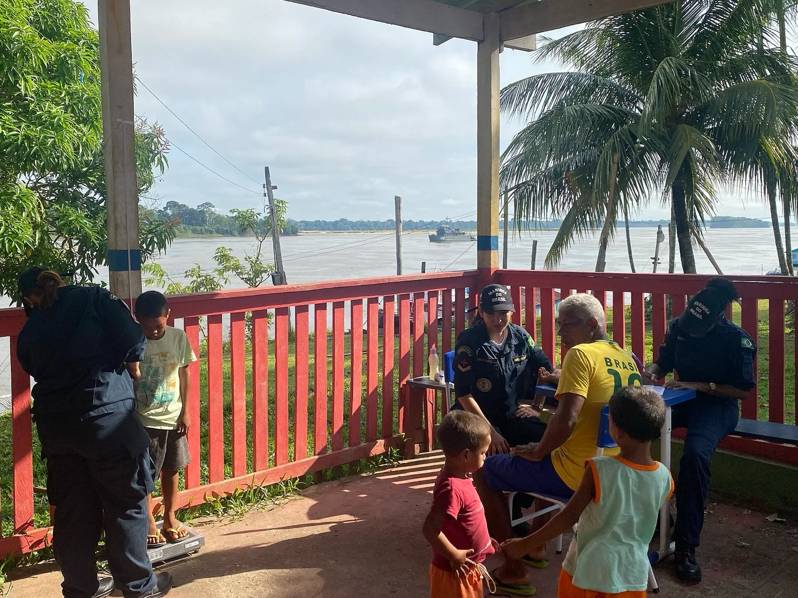 Militares da Marinha atendem moradores em uma varanda à beira de um rio, com um navio ao fundo, no Amazonas