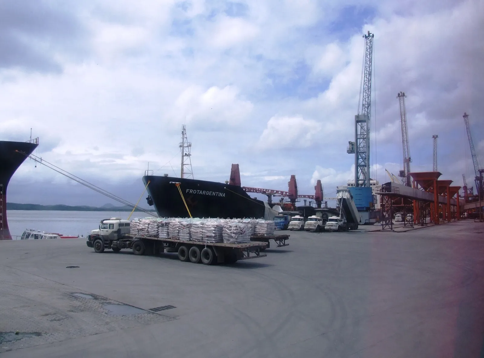 Caminhão carregado com sacas estacionado no cais, ao lado de navios graneleiros atracados e guindastes portuários