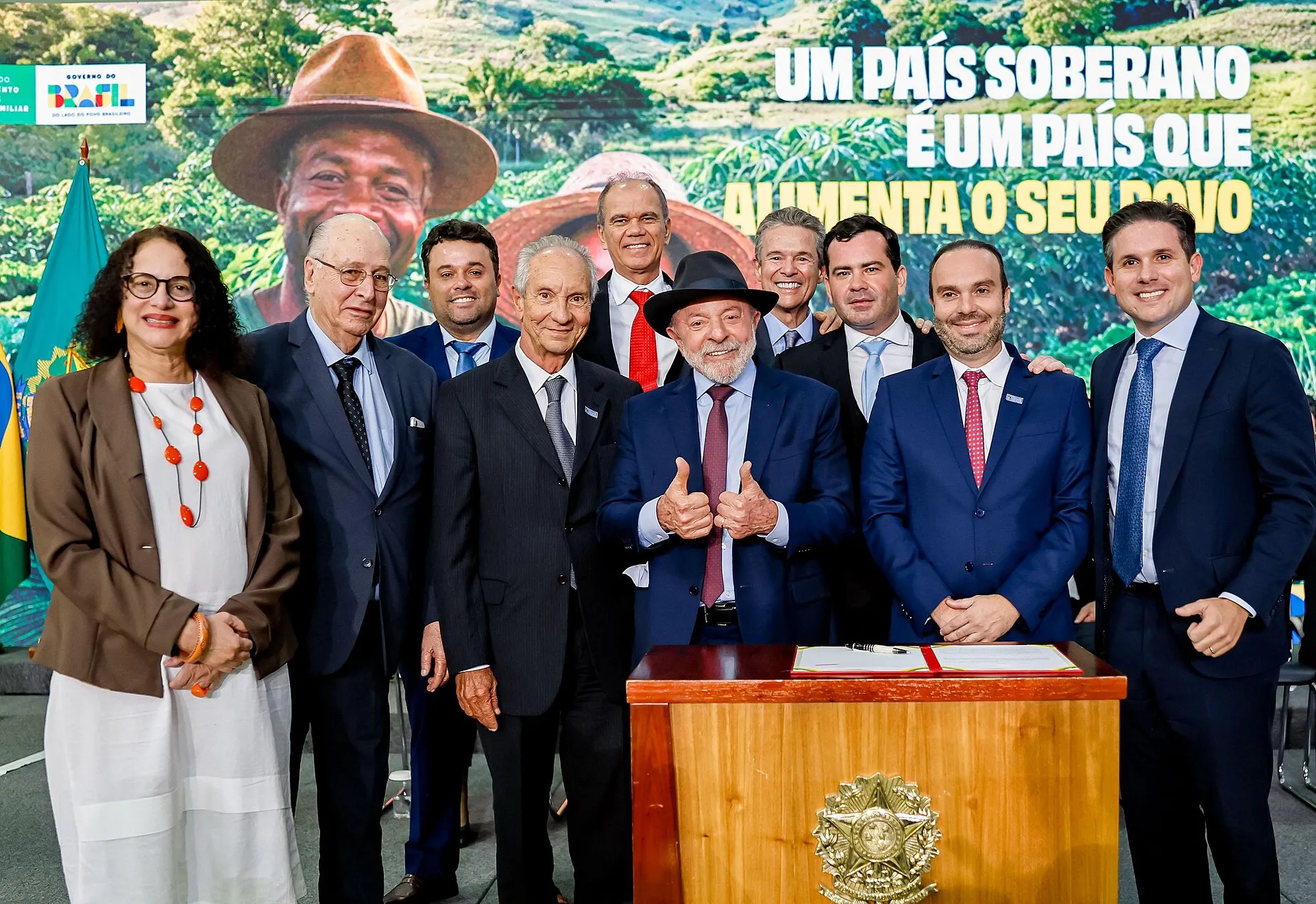 Grupo de autoridades posa atrás de uma mesa de madeira com o brasão da República, sob um painel com a frase 'Um país soberano é um país que alimenta o seu povo'