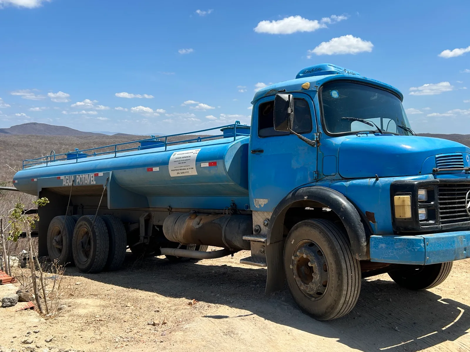 Caminhão-pipa azul com a faixa 'Água potável' e um adesivo da Operação Carro-Pipa, estacionado em paisagem seca do semiárido cearense